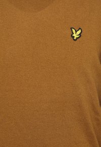 Brązowy dziergany sweter o prążkowanej fakturze z małym czarno-żółtym haftowanym logo orła na lewej piersi.