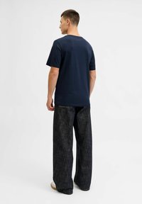 Marineblauw katoenen T-shirt met een ronde hals, gecombineerd met donkere denim wijde broek en lichtgekleurde schoenen. Achteraanzicht getoond.