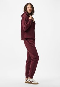 Γυναίκα που φοράει σετ μπορντώ φούτερ και φόρμες, κρατώντας τον γιακά του φούτερ, όρθια με άσπρα αθλητικά παπούτσια σε απλή φόντο.