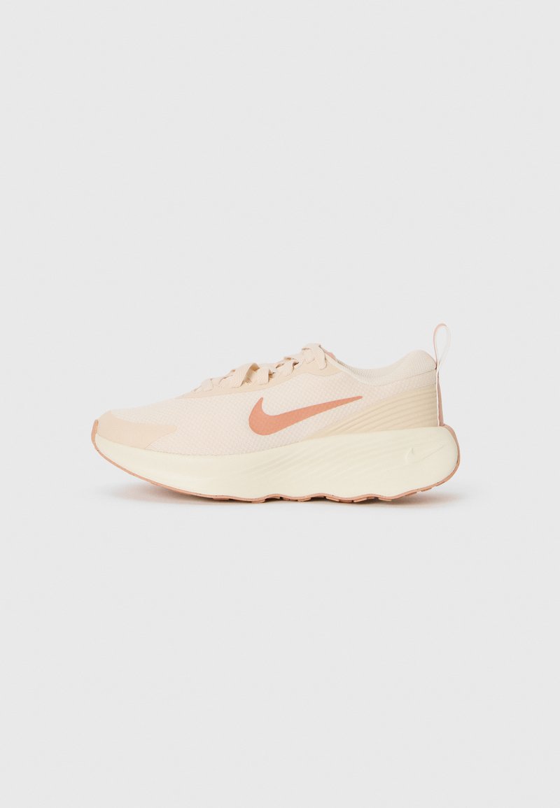 Helle beige Nike Laufschuhe mit einem pfirsichfarbenen Swoosh-Logo, dickem gepolstertem Sohlen und Zuglasche auf schlichtem weißen Hintergrund.