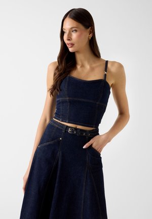 Femme aux longs cheveux bruns portant un débardeur court en denim bleu foncé et une jupe assortie avec ceinture, posant avec une main dans la poche.