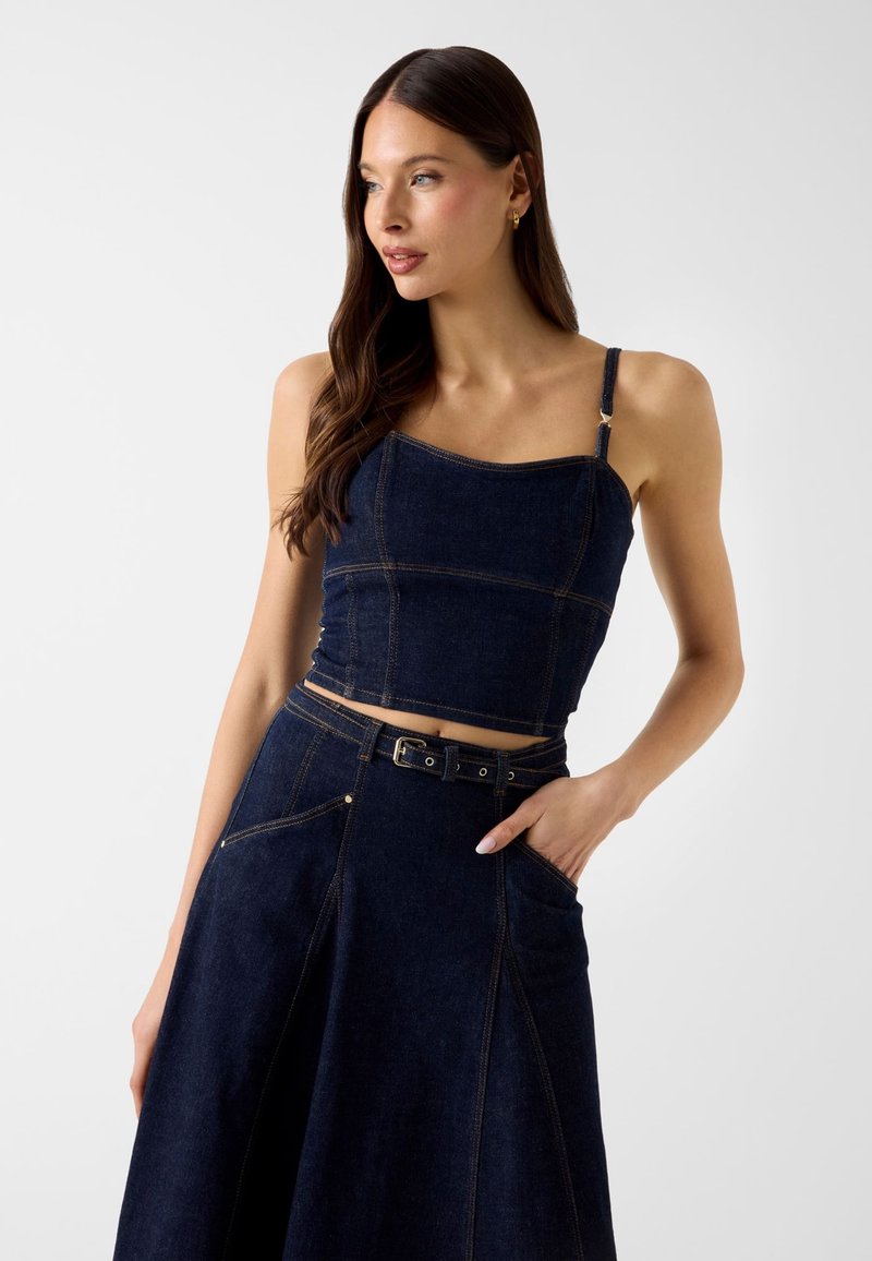 Femme aux longs cheveux bruns portant un débardeur court en denim bleu foncé et une jupe assortie avec ceinture, posant avec une main dans la poche.