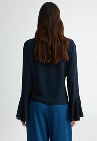 Námořnická modrá blůza s dlouhými, rozšířenými rukávy, vyrobená z hladkého materiálu. Zadní pohled ukazuje jednoduchý design bez ozdob.