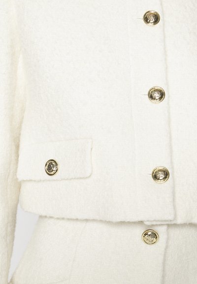 Λευκό υφασμένο μπουφάν με κοντό σχέδιο, χρυσά κουμπιά με λιοντάρι στο έμβλημα και μία μόνο τσέπη στο στήθος με αντίστοιχο χρυσό κουμπί.