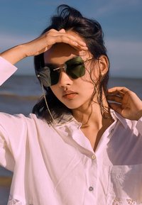 Grün getönte Sonnenbrille mit goldenen Akzenten, getragen mit einem weißen Hemd mit Knöpfen und Spitzenverzierungen. Der Hintergrund zeigt eine Strandlandschaft.