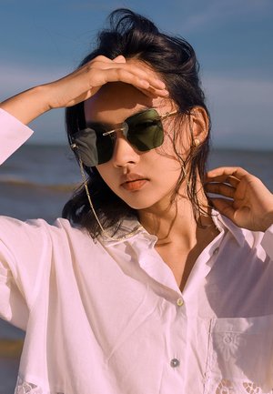 Grøntonede solbriller med guld detaljer, båret med en hvid skjorte med knap og blondedetaljer. Baggrunden viser et strandlandskab.