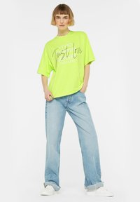 Neon-grünes, übergroßes T-Shirt mit bedrucktem Text, kombiniert mit hellblauen Schlaghosen und weißen Turnschuhen. Einfaches Design und lockerer Sitz.