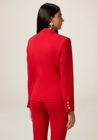 Czerwony marynarka skrojona z usztywnionego materiału, charakteryzująca się dopasowanym fasonem, detalami na szwie tylnym oraz guzikami na mankietach.