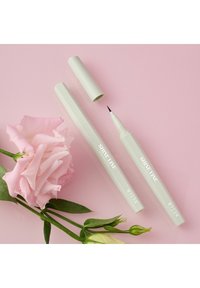 Penna per eyeliner bianco con punta fine, cappuccio rimosso, posizionata accanto a un bocciolo di rosa rosa e foglie verdi su uno sfondo rosa chiaro.