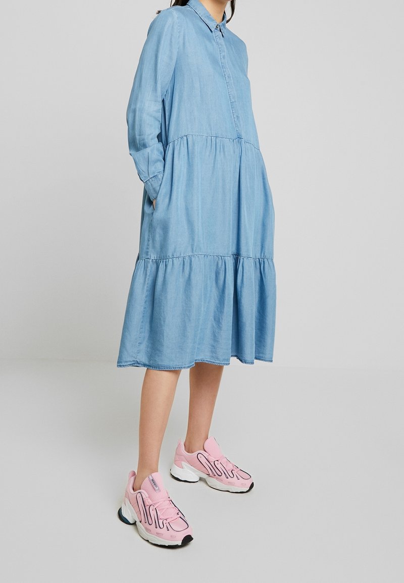 Femme portant une robe midi ample à manches longues bleu clair, associée à des baskets épaisses roses et blanches, debout les mains dans les poches.