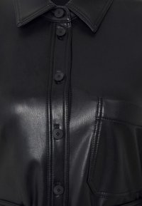 Černá kožená košile s límcem, která má přední zapínání na knoflíky a náprsní kapsu. Hladká textura s viditelnými detaily šití.