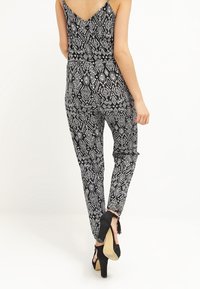 Mujer vestida con un mono sin mangas de patrón geométrico en blanco y negro y sandalias de tacón negro, alejándose sobre un fondo blanco.