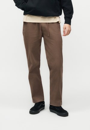 Homme portant un pantalon marron droit avec taille à cordon, baskets noires, sweat à capuche noir et chemise beige superposée en dessous.