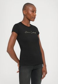 Musta puuvillapaita, jossa on lyhyet hihat ja kultaa brodeerattu "Armani Exchange" -logo keskellä rintaa.