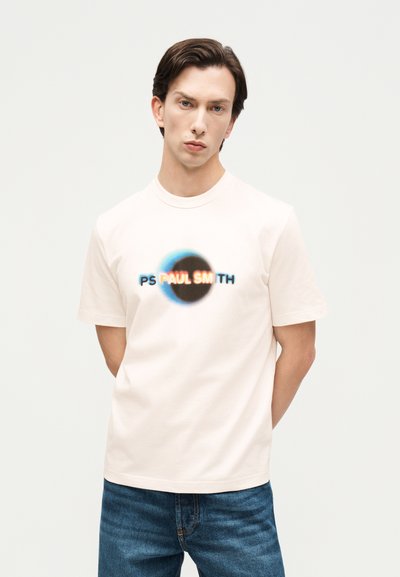 Mann iført en hvit t-skjorte med en uskarp svart sirkel og fargerik "PS PAUL SMITH"-tekst, kombinert med blå jeans, mot en ensfarget bakgrunn.