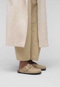 Sabots beiges avec une finition en daim, ornés d'une boucle en métal doré. Portés avec un pantalon large beige et un manteau de couleur claire.