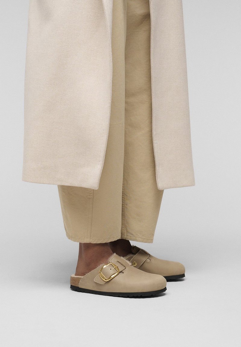 Sabots beiges avec une finition en daim, ornés d'une boucle en métal doré. Portés avec un pantalon large beige et un manteau de couleur claire.