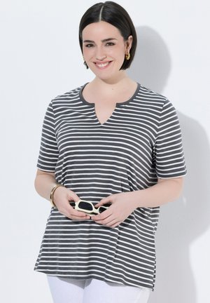 Lächelnde Frau mit kurzen dunklen Haaren, die ein grau-weiß gestreiftes kurzärmeliges Shirt trägt und weiße Sonnenbrillen hält, vor weißem Hintergrund.