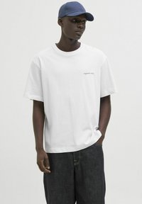 Camiseta de algodón blanca con mangas cortas, que presenta un pequeño logo en negro que dice "originals studio". Llevada con pantalones cortos de mezclilla oscura y una gorra azul.
