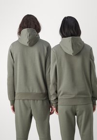 Sudaderas de color verde oliva con un corte relajado, que cuentan con un bolsillo tipo canguro y puños acanalados, combinadas con joggers a juego. Textura de tela suave.