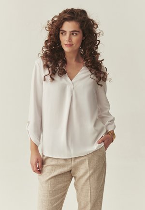 Femeie cu păr creț castaniu, purtând o bluză albă cu decolteu în V și pantaloni bej, stând cu o mână în buzunar, privind spre lateral.