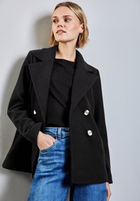 Abrigo peacoat de mezcla de lana negra con frente de doble botonadura, grandes botones dorados, cuello con muesca y corte relajado. Combinado con jeans azules.