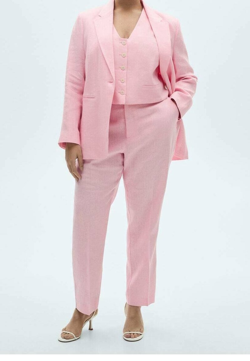 Femme portant un costume rose sur mesure avec gilet et pantalon assortis, debout avec une main dans la poche et chaussée de talons blancs ouverts à l'avant.