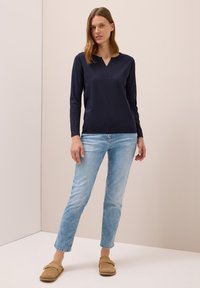 Marineblaues Langarmoberteil mit V-Ausschnitt, kombiniert mit hellblauen Slim-Fit-Jeans und beigen Loafers, getragen von einem Modell vor einem neutralen Hintergrund.