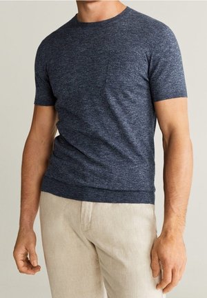 Man draagt een nauwsluitend, navyblauw gebreid shirt met korte mouwen en beige broek, staand met ontspannen armen tegen een effen achtergrond.