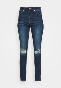 Jeans skinnies de talle alto en azul oscuro, con rodillas rasgadas y sutiles parches desgastados en los muslos, exhibidos sobre un fondo blanco.