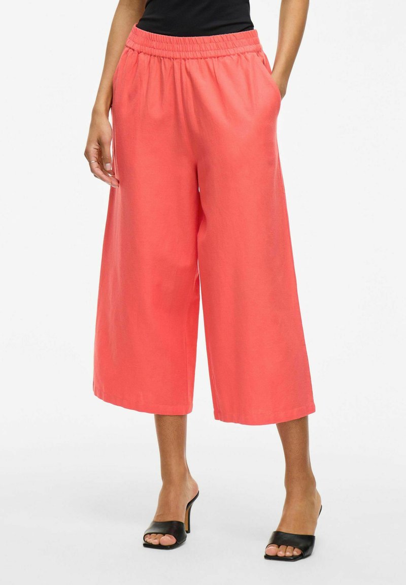 Pantalons larges en corail avec une taille élastique, dotés de poches latérales et d'une texture lisse, associés à des talons ouverts noirs.