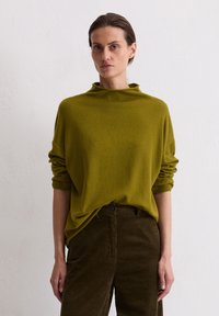 Olivgrüner, locker sitzender Strickpullover mit hohem Kragen und aufgerollten Ärmeln, kombiniert mit dunkelbraunen Cordhosen mit Knopfverschluss.