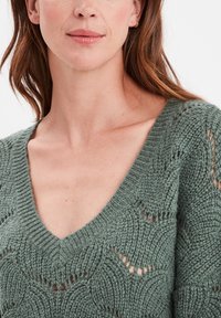 Pull en tricot de couleur verte douce, avec un décolleté en V profond et un motif complexe avec des mailles ouvertes, mettant en valeur la texture et les détails de conception.