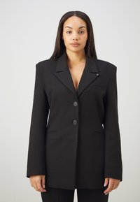 Blazer nero realizzato in tessuto texturizzato con rever a punta, taschino sul petto, due bottoni e un discreto dettaglio decorativo sul colletto.