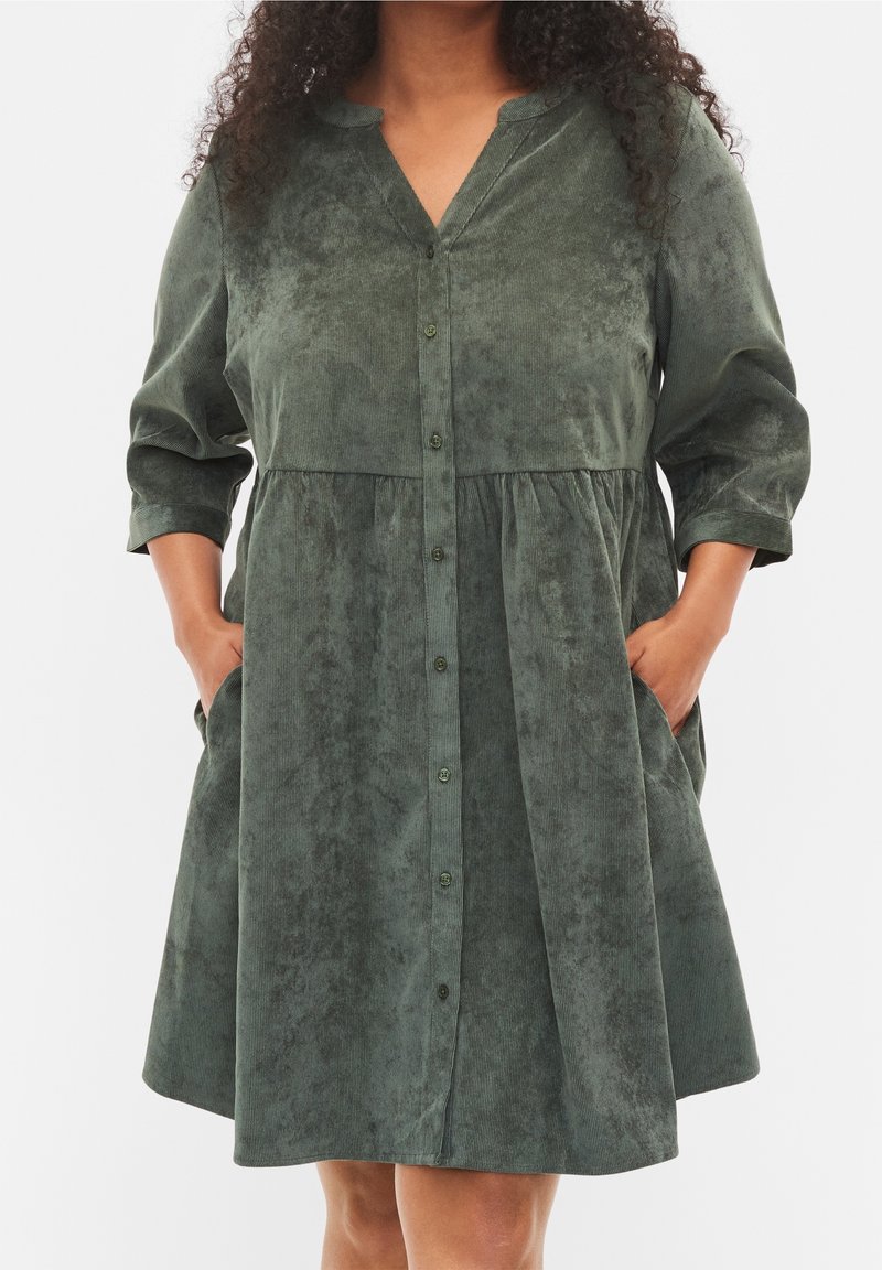 Femme portant une robe en velours côtelé vert foncé, ample et mi-longue, avec des boutons devant et les mains dans les poches latérales.
