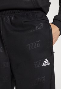 Černé kalhoty s jemným celoplošným vzorem loga Adidas, elastický pas a bílý akcent loga Adidas poblíž přední kapsy.