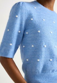 Maglione lavorato a maglia blu chiaro con maniche a palloncino corte, caratterizzato da dettagli in pompon bianchi e un orlo a costine, realizzato in un materiale morbido e strutturato.