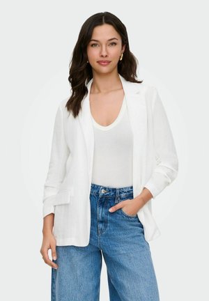 Femeie cu păr închis la culoare care poartă un sacou alb, top alb cu decolteu în V, blugi albaștri cu talie înaltă și cercei aurii, stând cu o mână în buzunar.