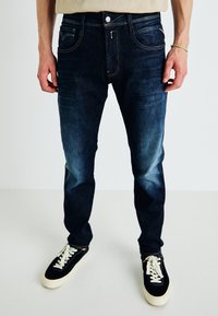 Mies, joka on pukeutunut tummansinisiin farkkuihin ja mustiin tennareihin, joissa on valkoiset nauhat, seisoo vaaleaa taustaa vasten.