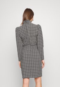 Rochie în carouri alb-negru cu guler înalt, mâneci evazate, talie ajustată și fustă până la genunchi. Material neted cu o croială structurată.