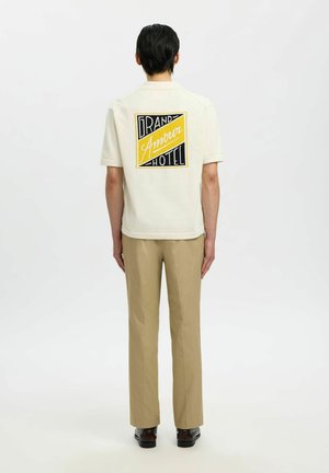 Mężczyzna ubrany w kremową koszulę z krótkim rękawem z naszywką z napisem „Grand Amour Hotel” na plecach, beżowe spodnie i ciemne buty, stojący tyłem.