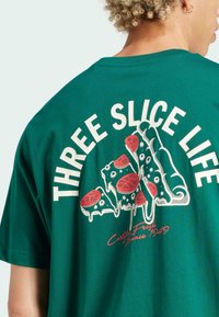 Vihreä puuvillapaita, jossa on pizzakuva grafiikalla "THREE SLICE LIFE" valkoisella ja punaisella, korostaen juuston ja pepperonin yksityiskohtia.