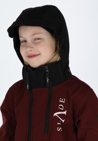 Red and black outerwear jacket with a high collar, front zippers, and a hood. Embroidered logo on left side. Smooth texture.