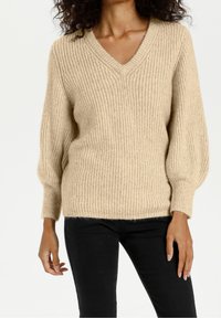 Pull beige en tricot avec encolure en V, texture côtelée et manches bouffantes. Porté avec un pantalon noir, affichant une coupe décontractée.