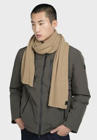 Beige ribbet tørklæde hængende om halsen, parret med en mørk grøn jakke med lynlås, der har lynlås-lommer og sorte detaljer.