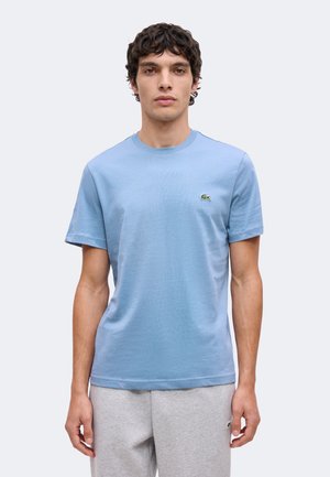 Joven con pelo corto y rizado que lleva una camiseta de manga corta azul claro y pantalones deportivos gris claro sobre un fondo liso.