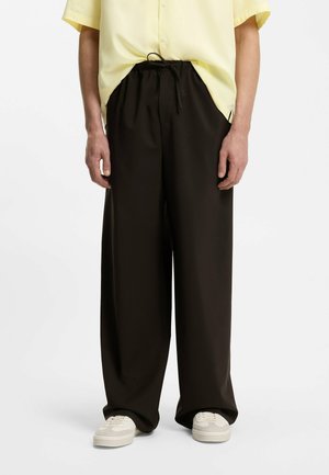 Mężczyzna w luźnych czarnych spodniach ze sznurkiem, żółtej koszuli z krótkim rękawem i białych tenisówkach, stojący na białym tle.