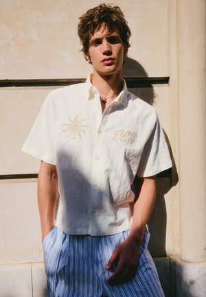 Young man with tousled hair wearing a white short-sleeve shirt with sun and "146" embroidery, and blue striped pants, standing against a beige wall.