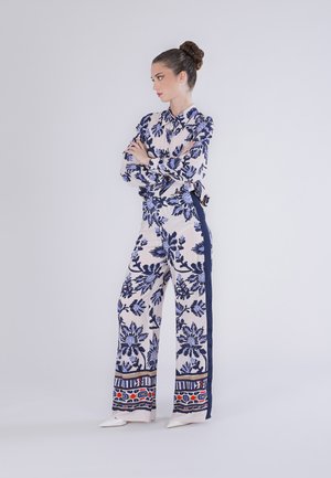 Mujer con el cabello recogido en un moño, vestida con blusa y pantalones de pierna ancha con estampado floral a juego, de pie con los brazos cruzados, mirando hacia un lado.