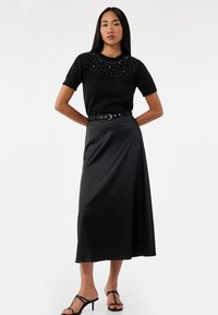 Svart stickad topp med nitar vid halsen, i kombination med en svart satin midi-kjol, åtsmitande vid midjan med ett bälte och hårdvaruaccenter.